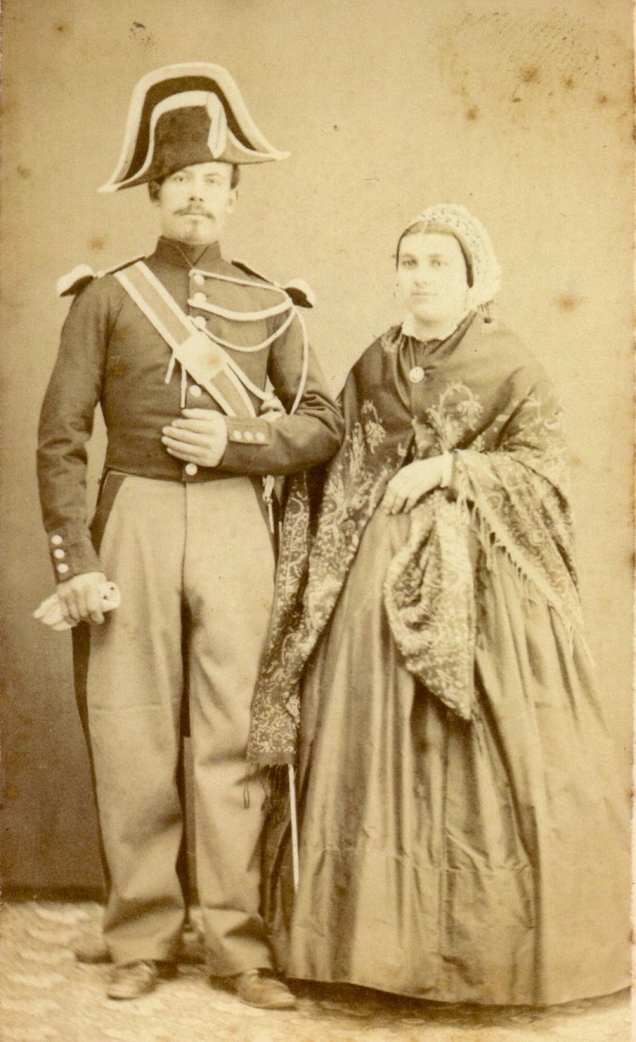 Portrait double. Il s’agit d’un gendarme en costume d’apparat et de son épouse. La présence militaire est importante dans une ville frontalière ou de nombreuses garnisons occupent les casernes (Saint-Martin, caserne du Puig, Citadelle). Son épouse porte sur les épaules un magnifique châle carré en laine cachemire, probablement fabriqué à Nîmes. La coiffe est parée de riches dentelles et rubans. Le châle est arrêté par une broche ainsi qu’une épingle. La crinoline est ronde. Il faut noter pour ce costume l’absence de tablier d’apparat. Photographie Scanagatti, Perpignan, vers 1865, collection Institut du Grenat.