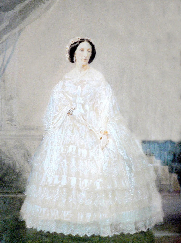 Portrait de la Comtesse Charlotte de LAZERME (1822-1888). Son haut rang lui permet de se montrer avec une vaporeuse crinoline. On perçoit à ses poignets d’imposants bracelets en or et pierreries. Son visage est mis en valeur par les boucles d’oreilles et la coiffe à la mode, ornementée de fleurs. Une simple chaîne est suspendue à son cou. Perpignan trouve en Charlotte de LAZERME l’une de ses égéries de mode. Sa fortune en fait une femme du monde, position propice à devenir un modèle d’élégance et de nouveauté pour les femmes du Roussillon. Aquarelle, vers 1860, Perpignan, collection particulière.